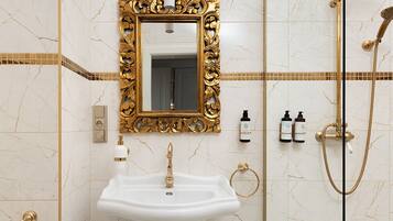 Casa de banho com lavatório pedestal branco, espelho com moldura dourada, chuveiro dourado e sanita.