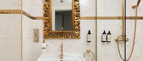 A bathroom with a white pedestal sink, a gold-framed mirror, a gold showerhead, and a toilet.