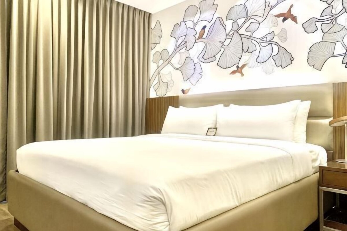 A hotel room with a large bed, a headboard with a light fixture, a nightstand with a lamp, and a wall with a floral and bird mural.