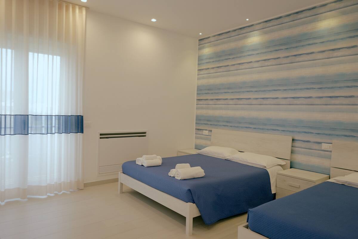 Moderni hotellihuone, jossa on kaksi sänkyä, suuri ikkuna läpikuultavilla verhoilla ja kuvioitu seinä.