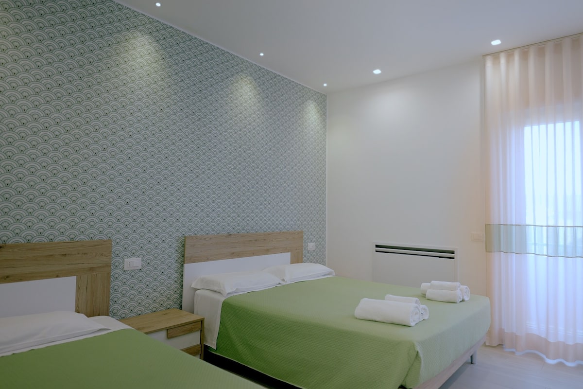 Hotellihuone, jossa on kaksi sänkyä, vihreä päiväpeite, kuvioitu tapetti ja ikkuna, jossa on läpikuultavat verhot.