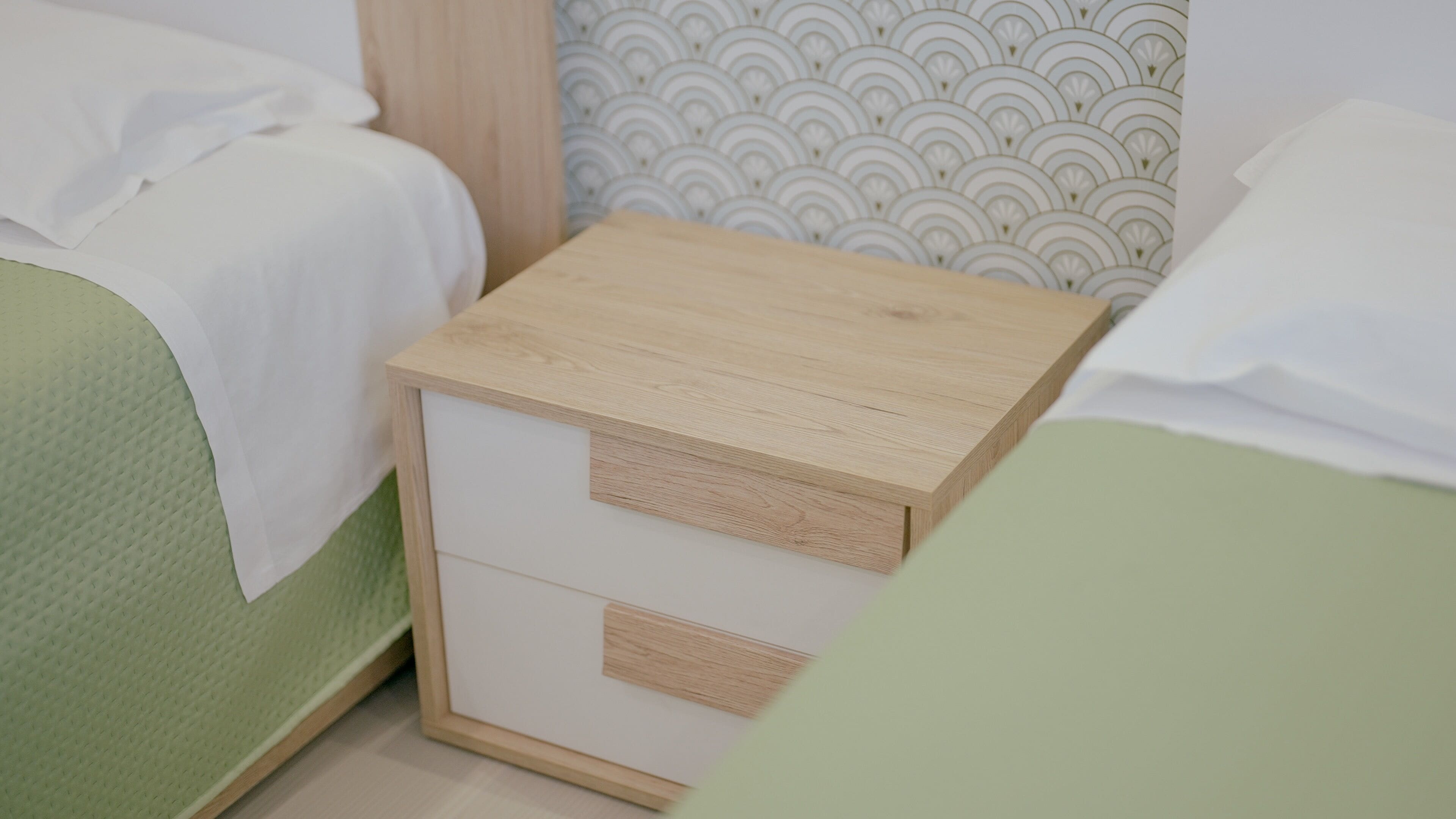 A small wooden bedside table with a drawer, positioned between two beds with green bedding.