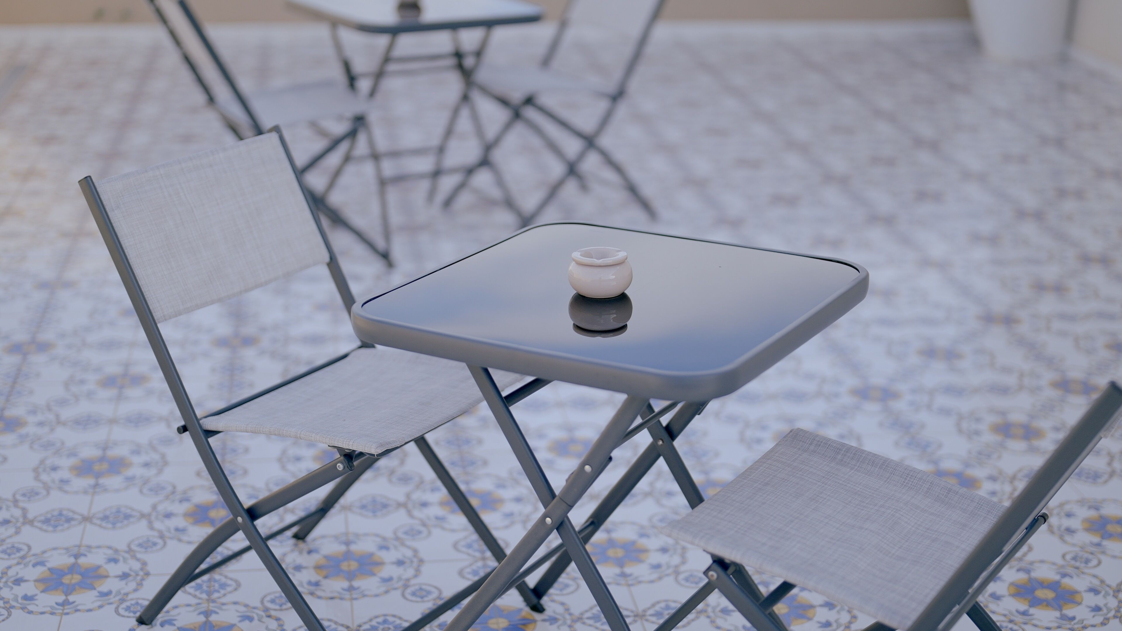 Une petite table carrée sur laquelle se trouve un objet blanc, entourée de deux chaises dont les dossiers sont en treillis.