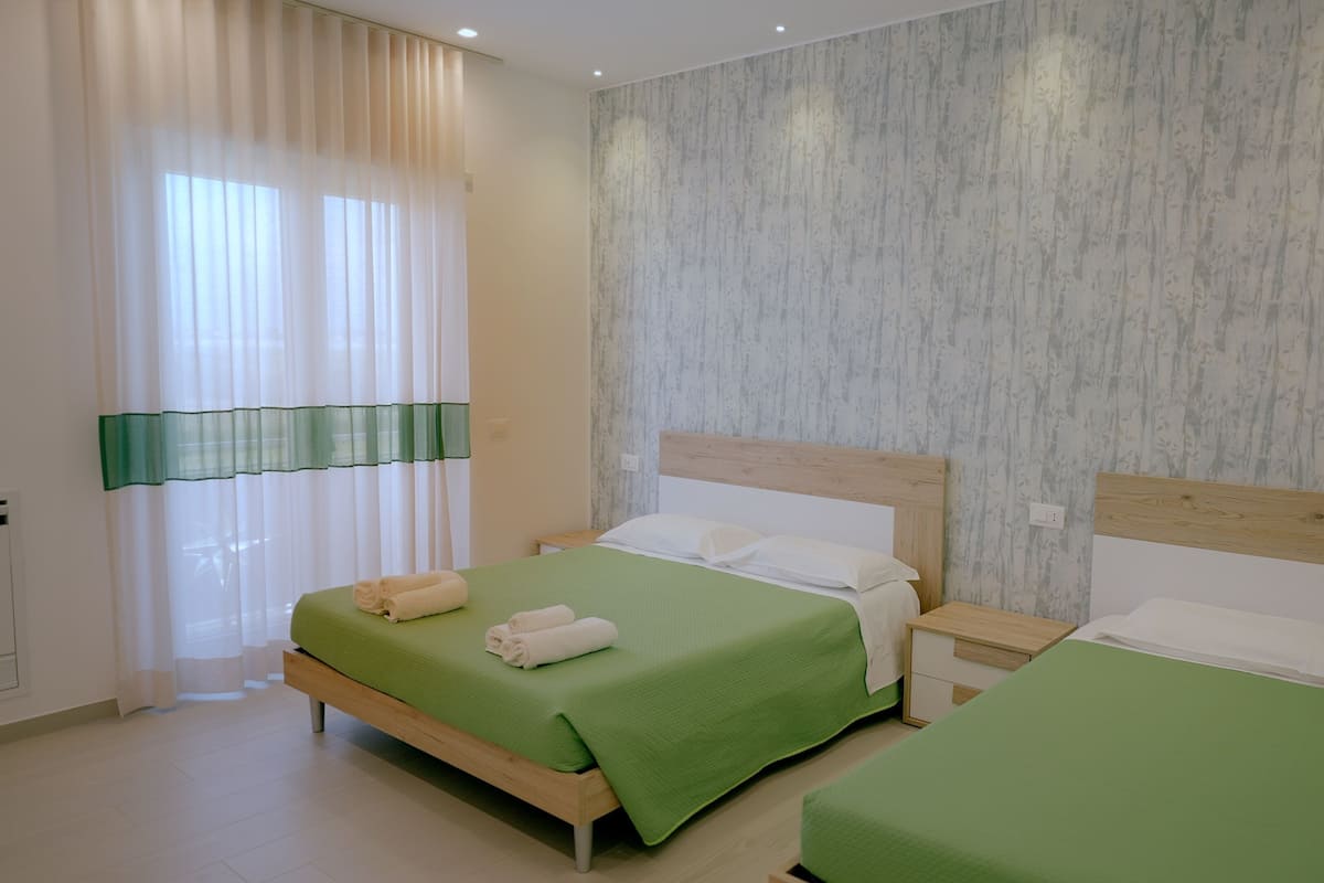 Hotellihuone, jossa on kaksi sänkyä, puinen pääty, vihreä peitto, valkoinen tyyny ja ikkuna, jossa on läpikuultavat verhot.