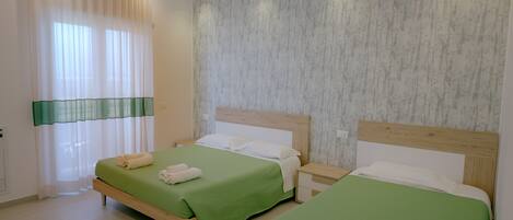 A hotel room with two beds, a wooden headboard, a green bedspread, a white pillow, and a window with sheer curtains.