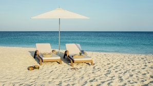 Two wicker lounge chairs with white cushions and a white umbrella on a sandy beach.