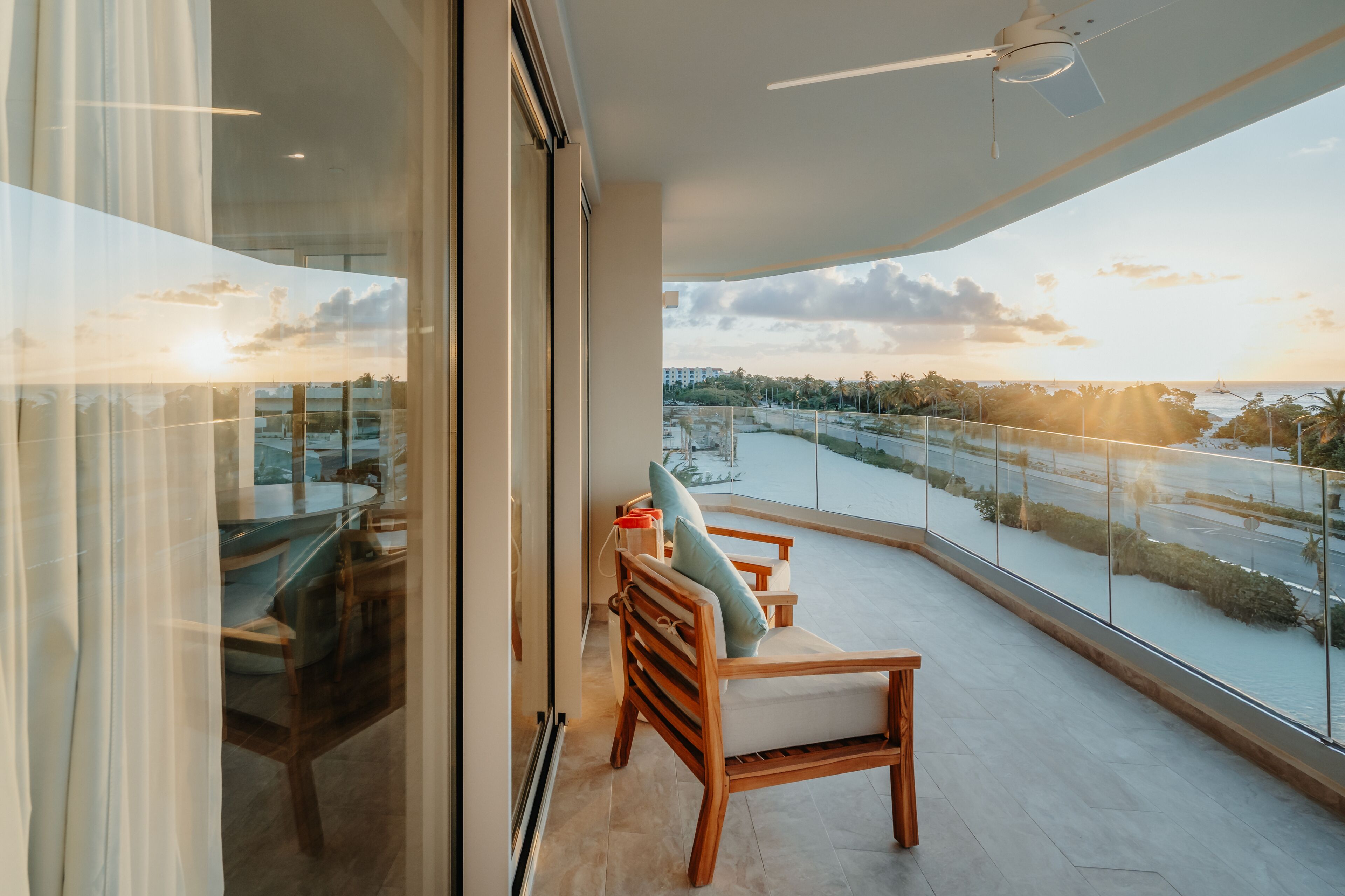 Ein Balkon mit Blick auf ein Gewässer und Palmen, ein Glastisch mit Stühlen sowie ein Deckenventilator.