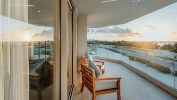 Ein Balkon mit Blick auf ein Gewässer und Palmen, ein Glastisch mit Stühlen sowie ein Deckenventilator.