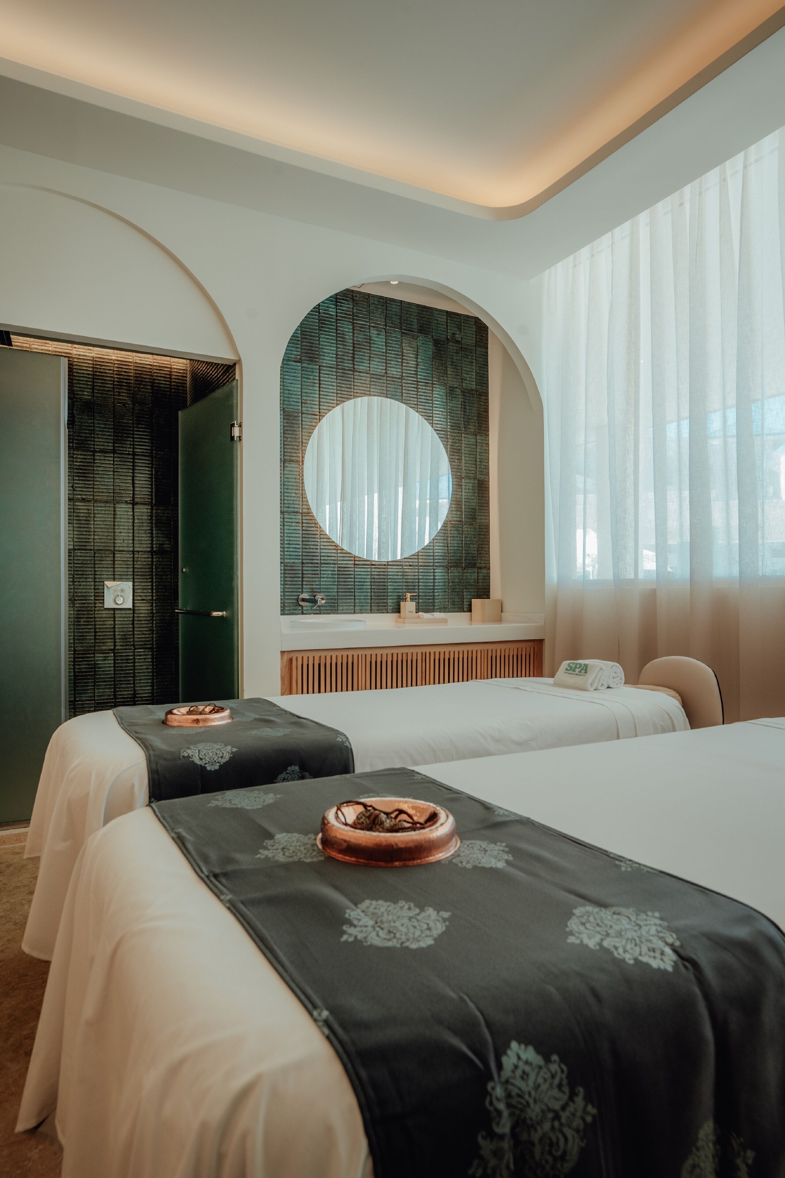 A hotel room with two beds, a round mirror, a vanity with a sink, and a large window with sheer curtains.