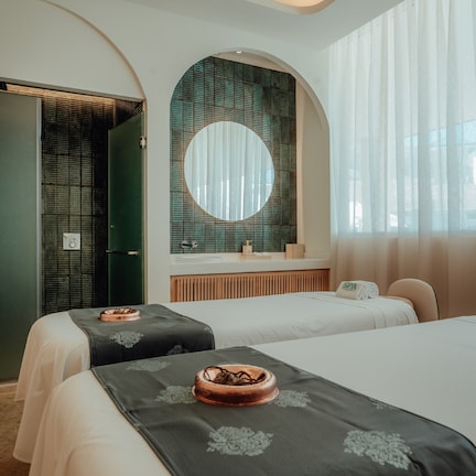 A hotel room with two beds, a round mirror, a vanity with a sink, and a large window with sheer curtains.