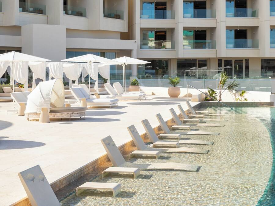 Ein Poolbereich mit Liegestühlen und Sonnenschirmen, ein klares Schwimmbecken und ein mehrstöckiges Gebäude mit Balkonen.