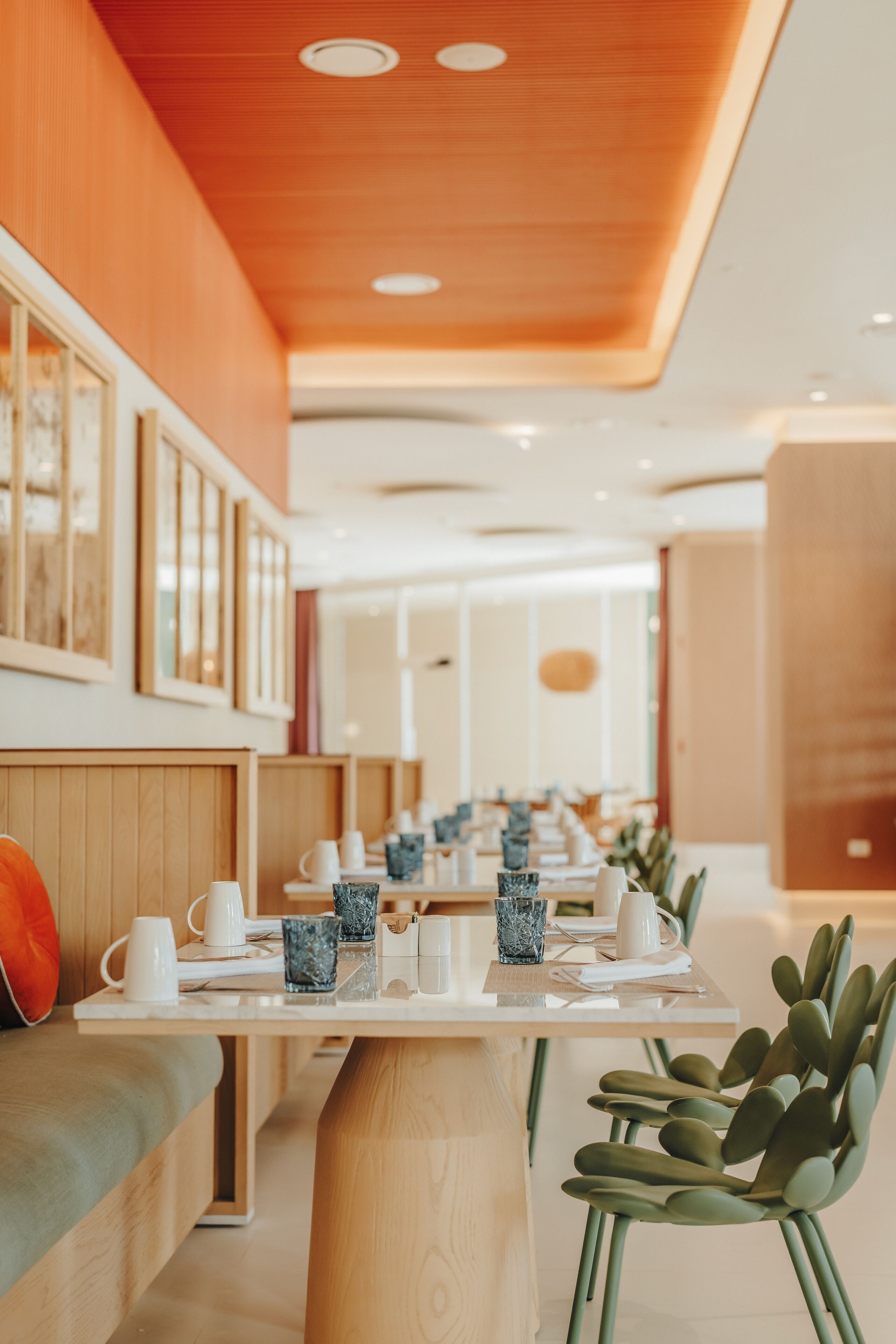 A modern dining area with wooden tables and green chairs, framed artwork on the walls, and a warm ceiling design.