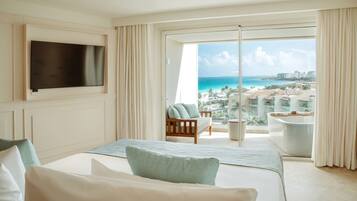 Habitación de hotel con cama, televisor y balcón con vista al océano y edificios.