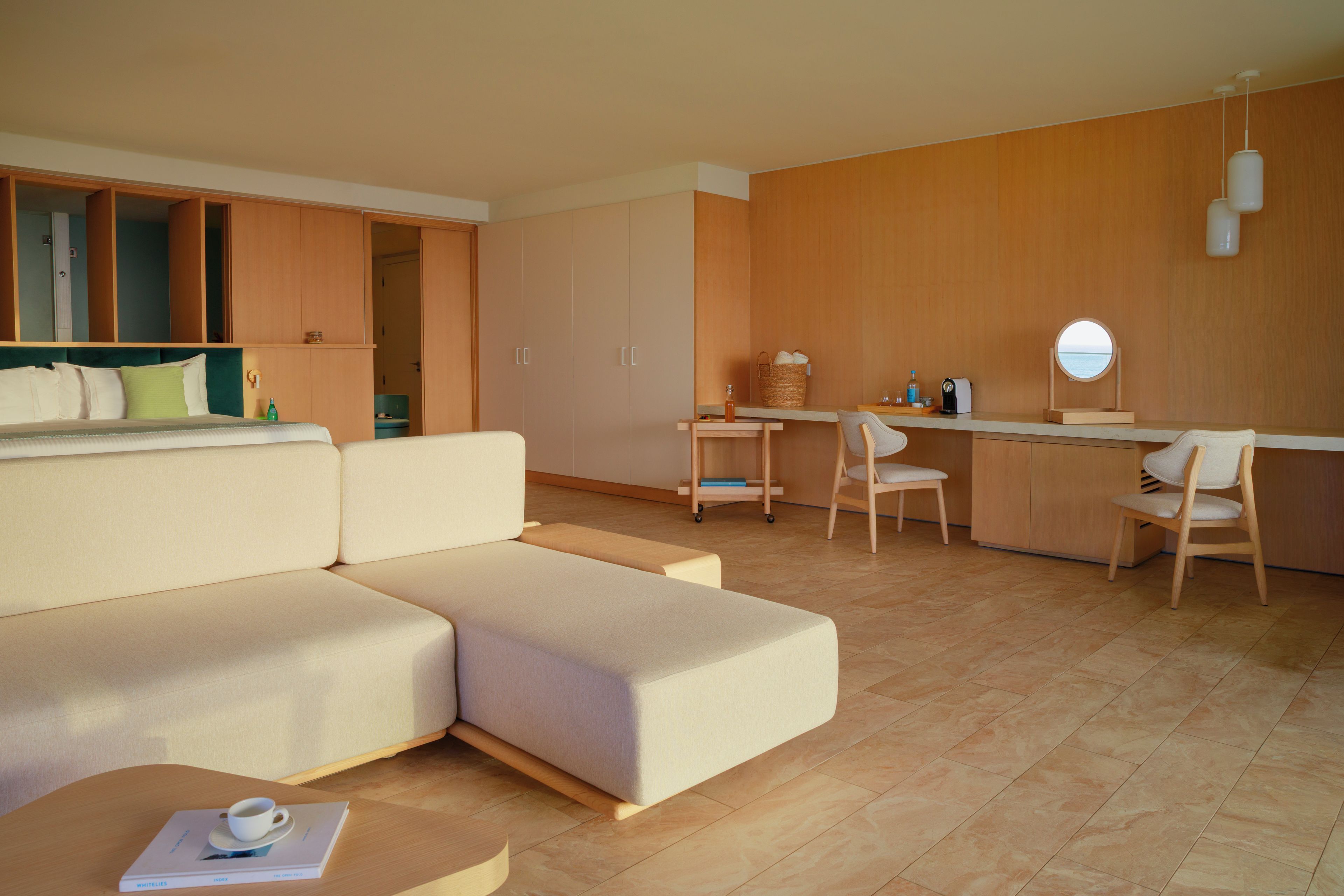 Habitación de hotel moderna con sofá, una pequeña mesa, un escritorio y una silla. Se observa un armario y un espejo en la pared.