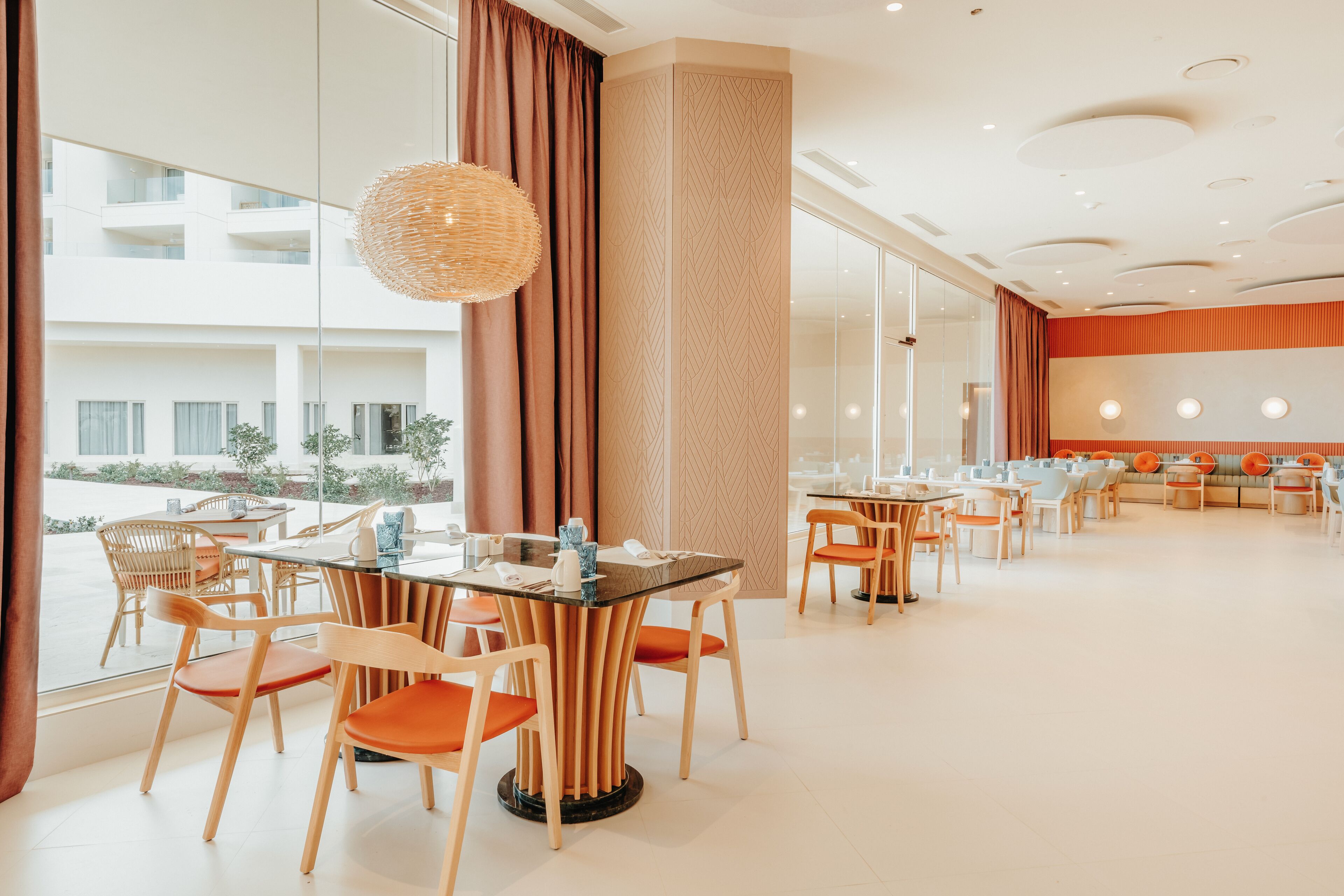 A modern dining area with wooden chairs and tables, large windows, and a view of the building's exterior.