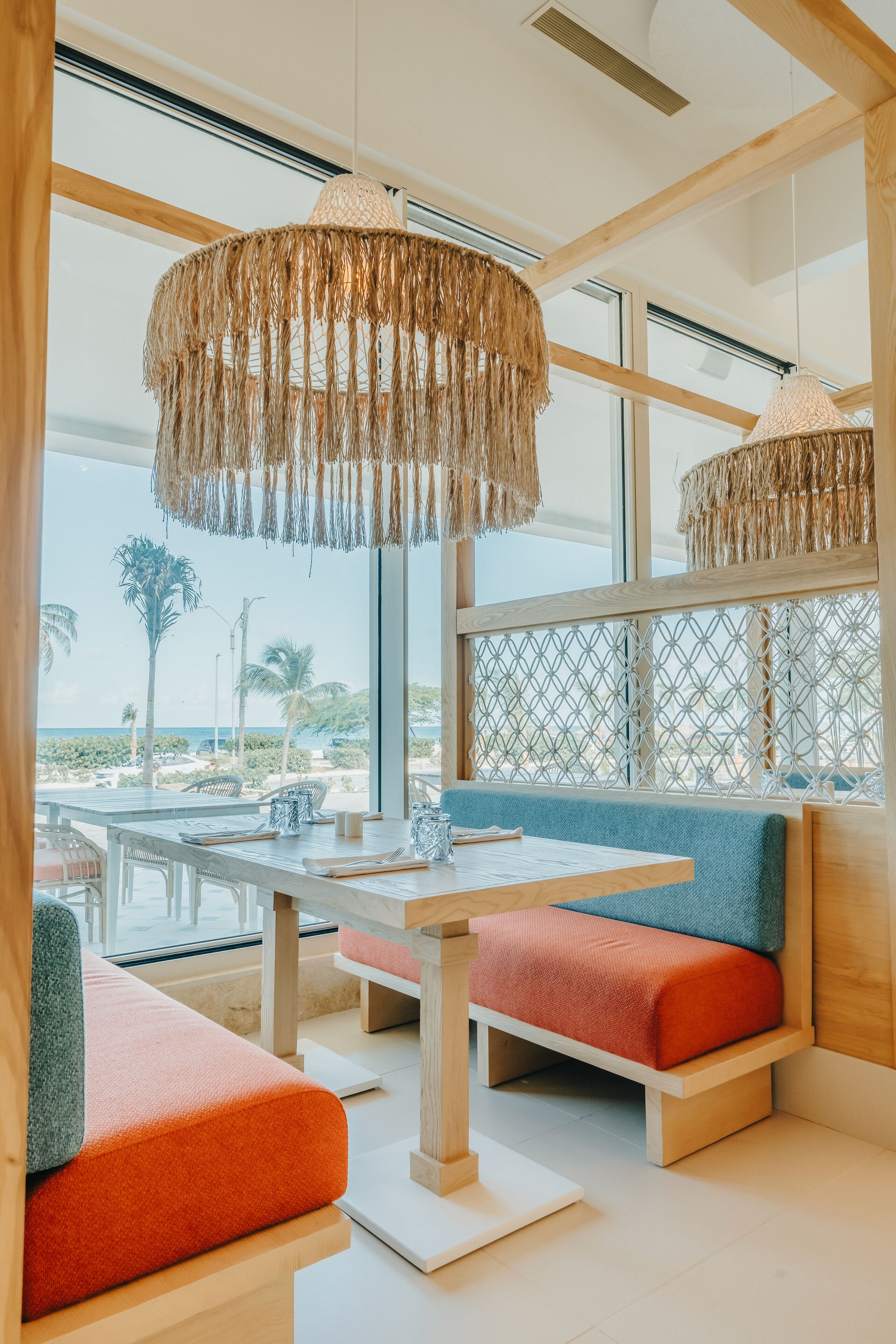 A modern interior with a wooden table, colorful seating, and large windows offering a view of the beach.