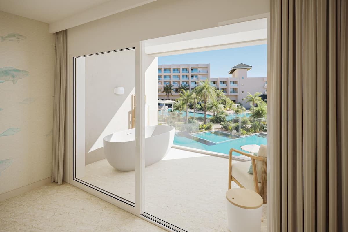 Et moderne hotelværelse med en stor skydedør, der fører til et poolområde, med et hvidt badekar og et lille bord med stole.