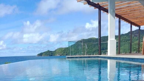 A clear blue infinity pool with a view of the ocean and a hilly landscape. - Lagati Residence (Mahé Island)