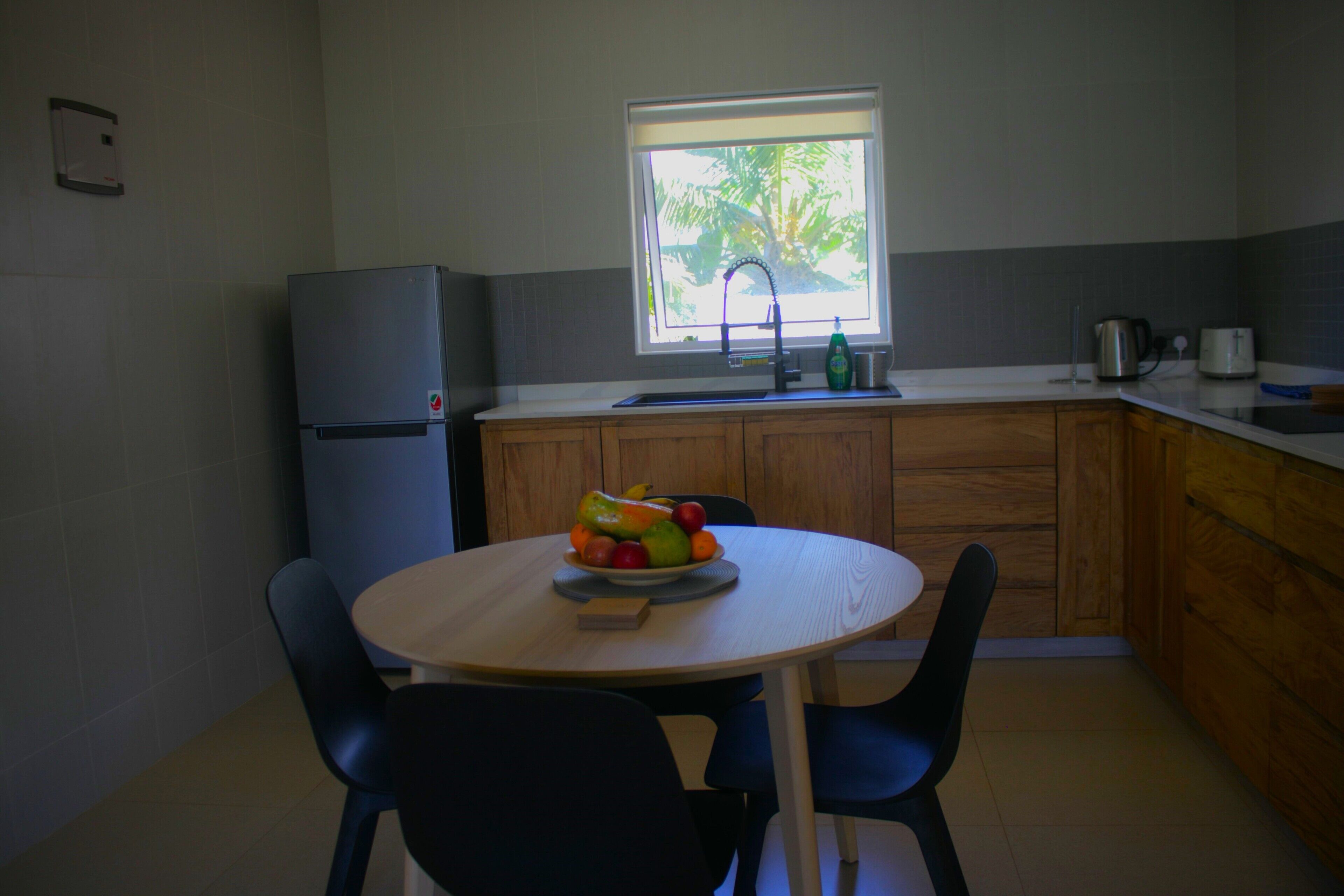 A compact kitchen with a round table, two chairs, a refrigerator, a window with a view of trees, and a sink.