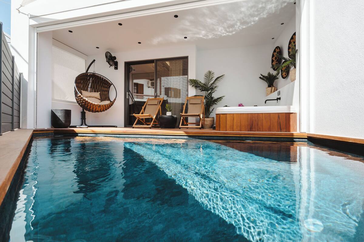 A modern indoor pool area with a clear blue pool, wooden deck, and a seating area with chairs and a hanging chair.