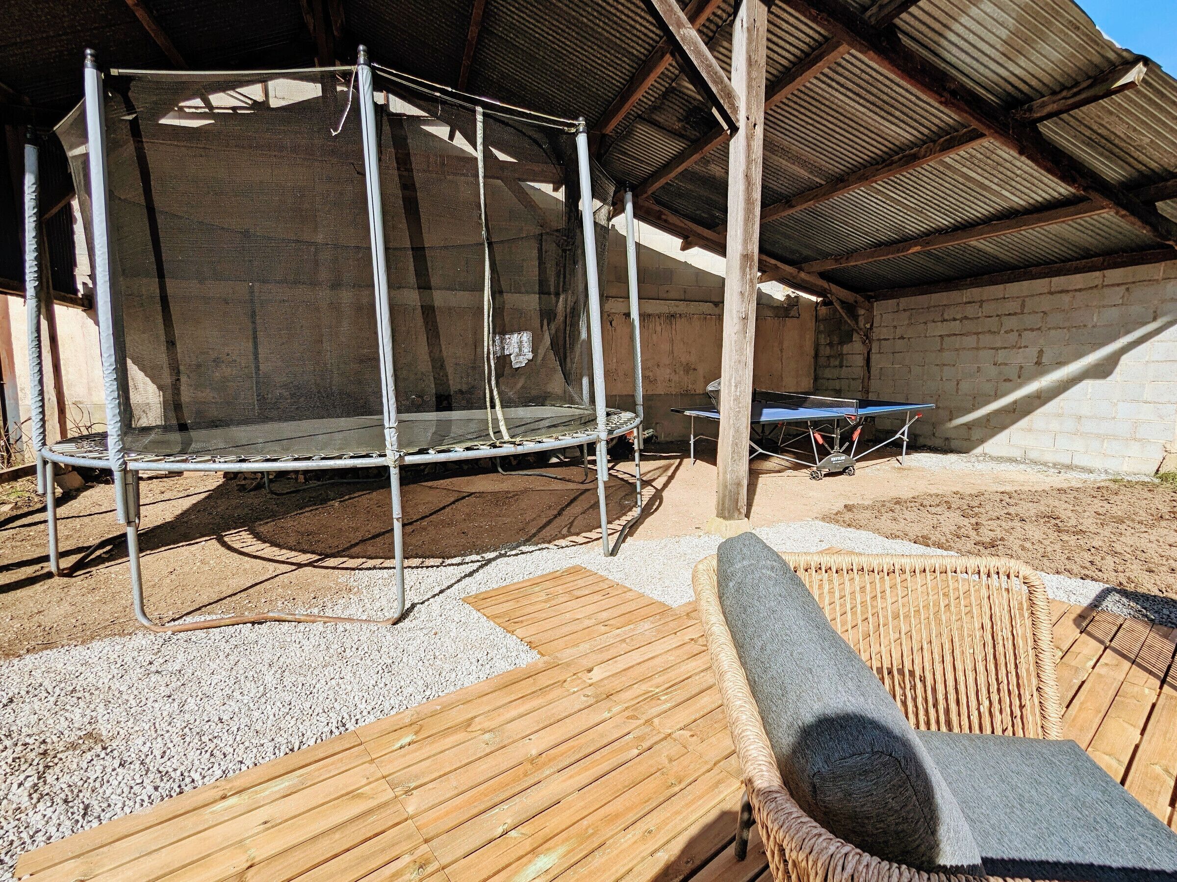 Un espace extérieur abrité comprenant un trampoline, une table de ping-pong et un fauteuil de relaxation en osier.
