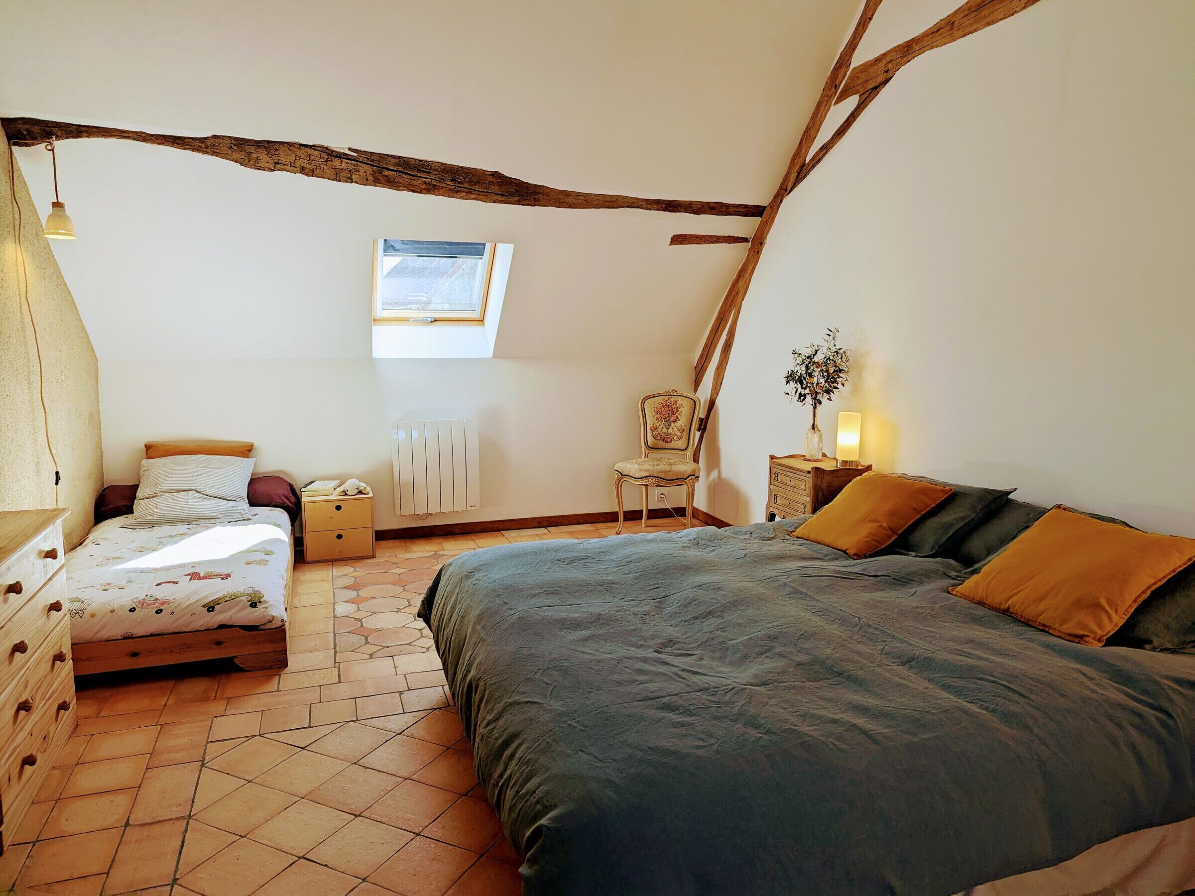 Une chambre avec un plafond en pente, un lit recouvert d’une couverture bleue, une chaise, une table de chevet avec une lampe et un radiateur.