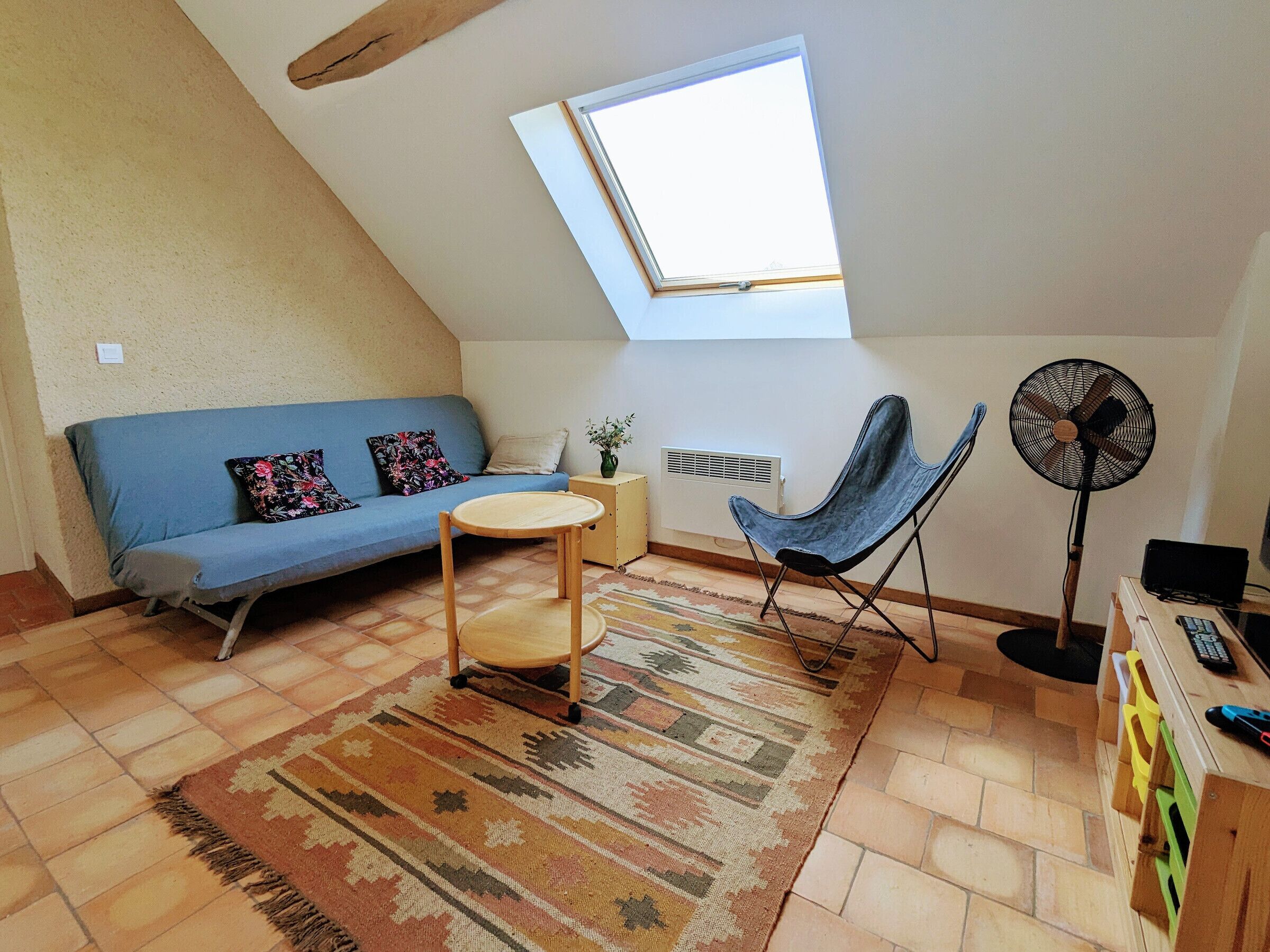 Une pièce confortable de style loft, avec un plafond en pente, un canapé bleu, une petite table ronde, une chaise pliante bleue, un ventilateur sur pied et un radiateur.
