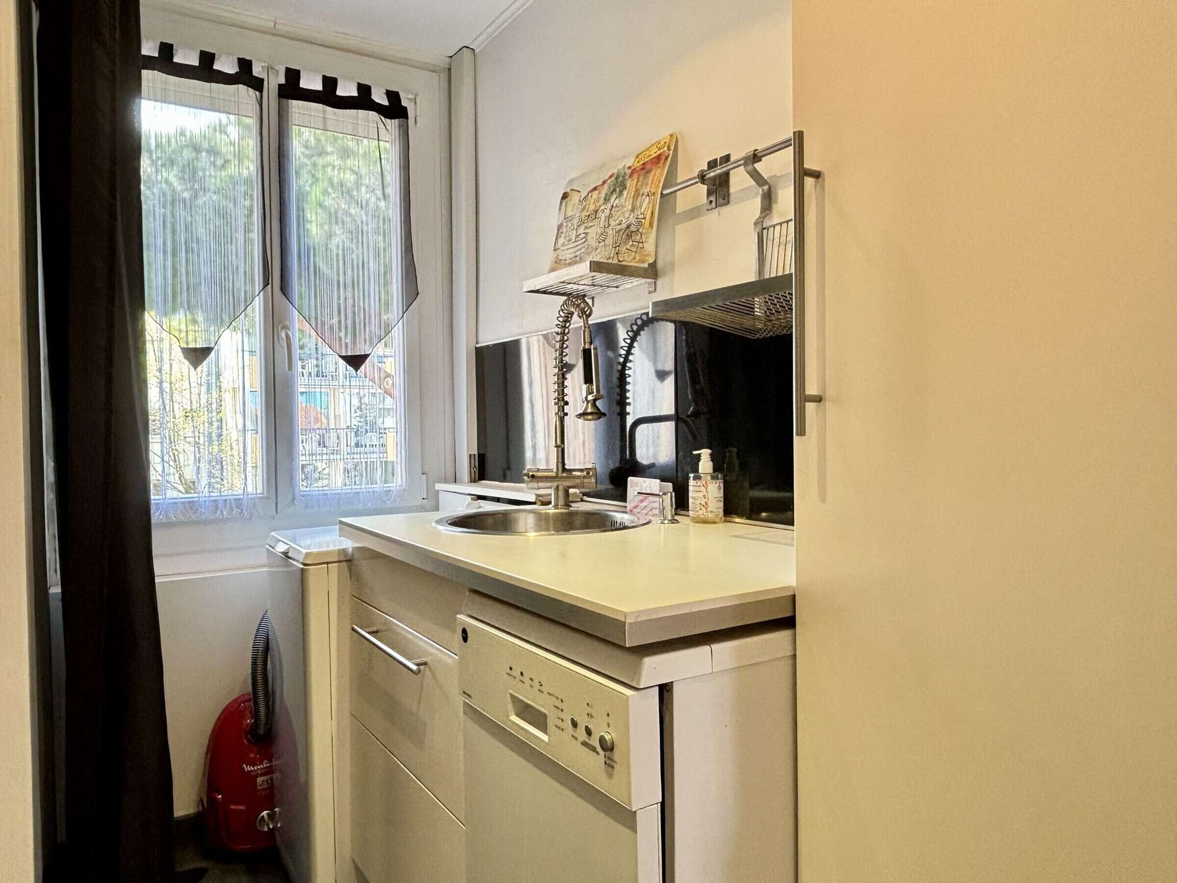 A compact kitchen with a sink, a dish rack, and a window with curtains.
