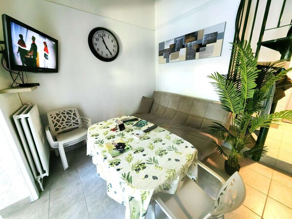 A cozy living area with a sofa, a table with a floral tablecloth, a clock, a television, and a plant. - Semi-detached villa with pool and parking for 4 people (Cap d'Agde)