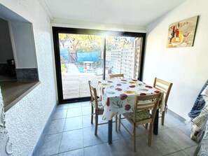 A dining area with a table and chairs, a view of the backyard through a sliding glass door, and a painting on the wall. - Semi-detached villa with pool and parking for 4 people (Cap d'Agde)