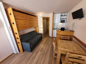 A compact room with a sofa, wooden table, and a kitchenette area.