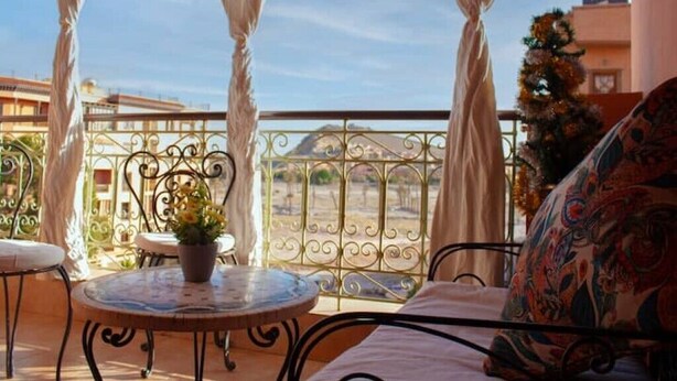 A balcony with a round table and chairs, a potted plant, and a view of a building labeled "MARRAKECH HOUSE".