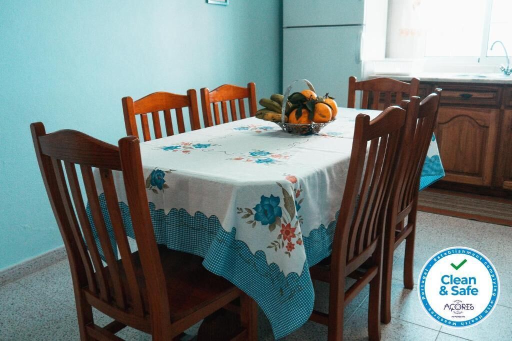 Una mesa de comedor con mantel floral, rodeada de sillas de madera. Hay una cesta de frutas sobre la mesa.