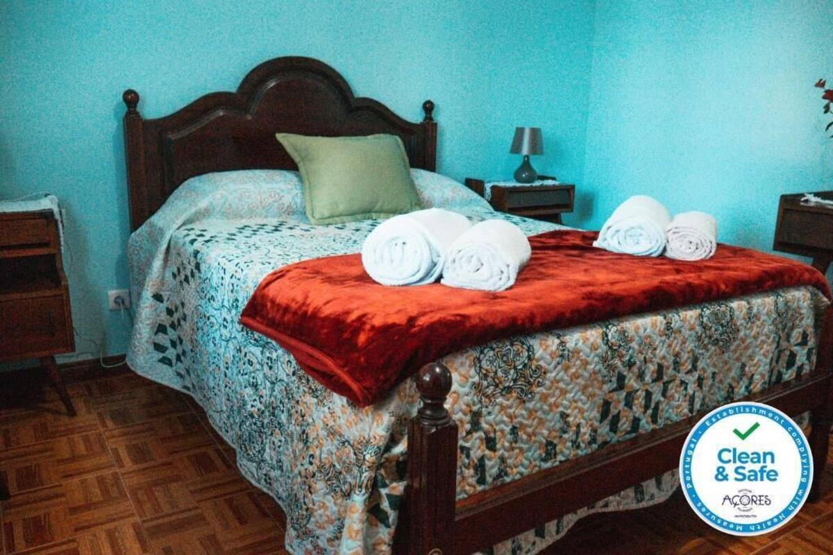 A neatly made bed with a red blanket, a green pillow, and rolled towels on the side.