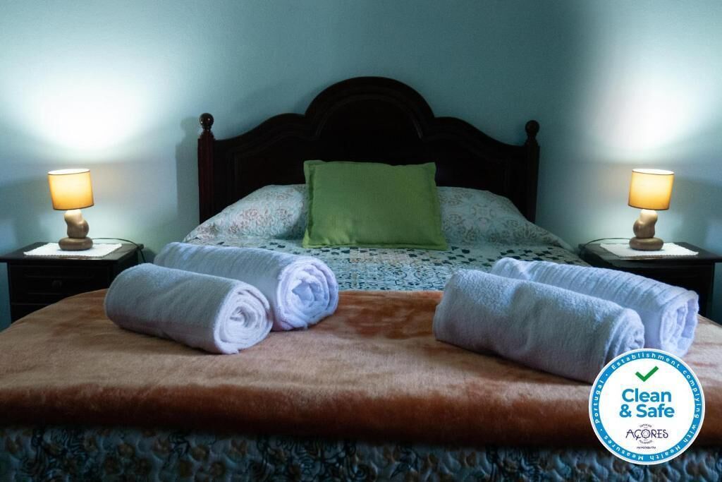 Una cama bien hecha con dos toallas blancas enrolladas, una almohada verde, dos lámparas de noche y un logotipo que indica que el establecimiento es limpio y seguro.