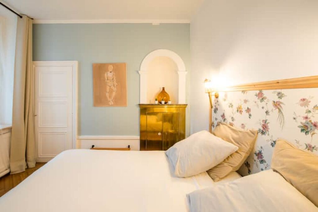 A bedroom with a bed, a nightstand, a wall-mounted lamp, and a floral wallpaper.