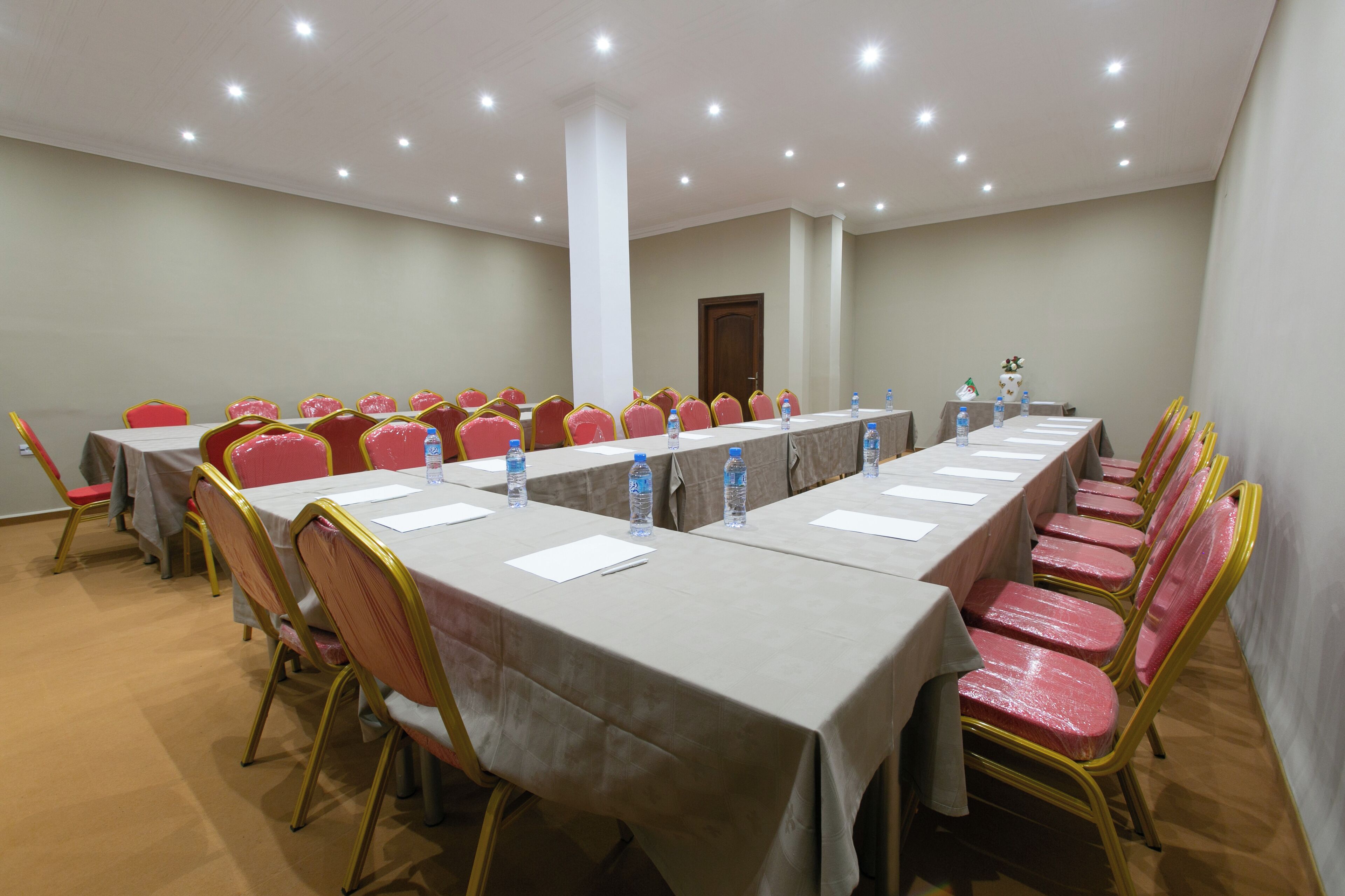Un salón de conferencias con una larga mesa, sillas rojas y botellas de agua.