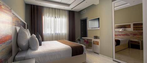 A modern hotel room with a large bed, a desk, a mirror, and a view of another room.