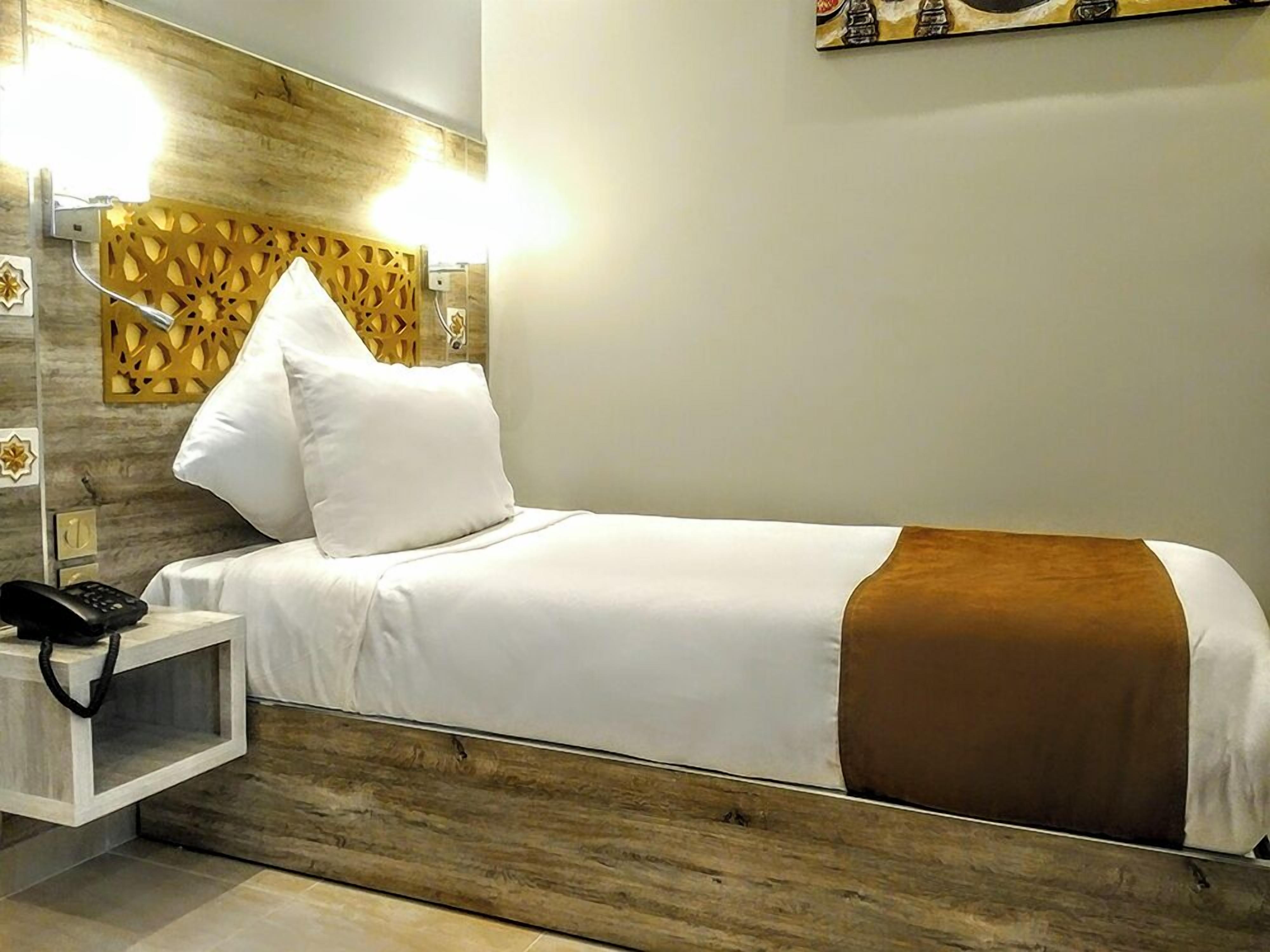 A neatly made bed with white bedding and a brown throw, a bedside table with a phone, and a wall-mounted light fixture.