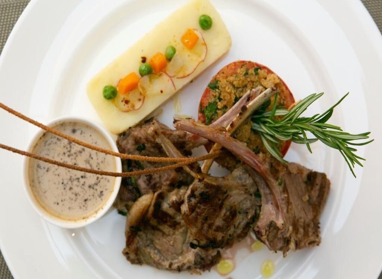 Un plato con chuletas de cordero, una rodaja de patata, guisantes, zanahorias y una ramita de romero.