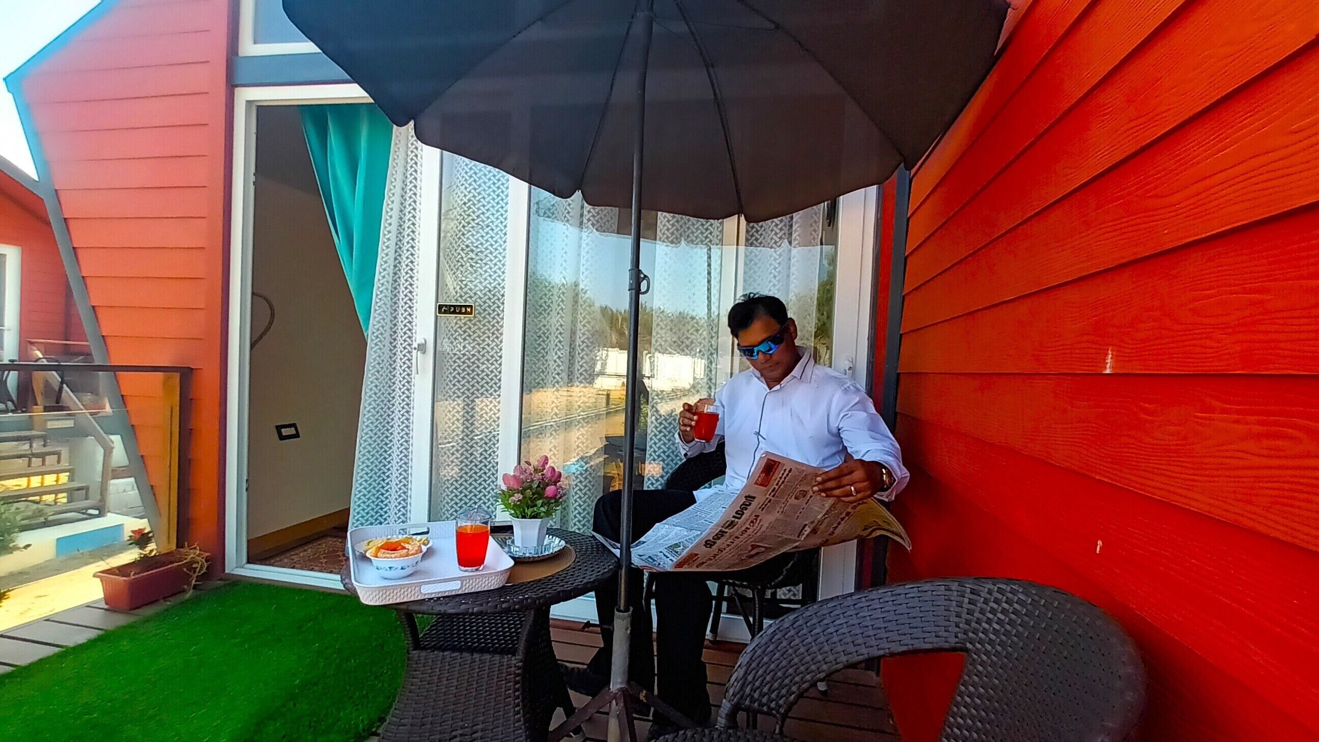 Una persona sentada a una mesa con un periódico, una bebida y un plato de comida en un patio con una puerta corrediza de cristal, una mesita y una silla.