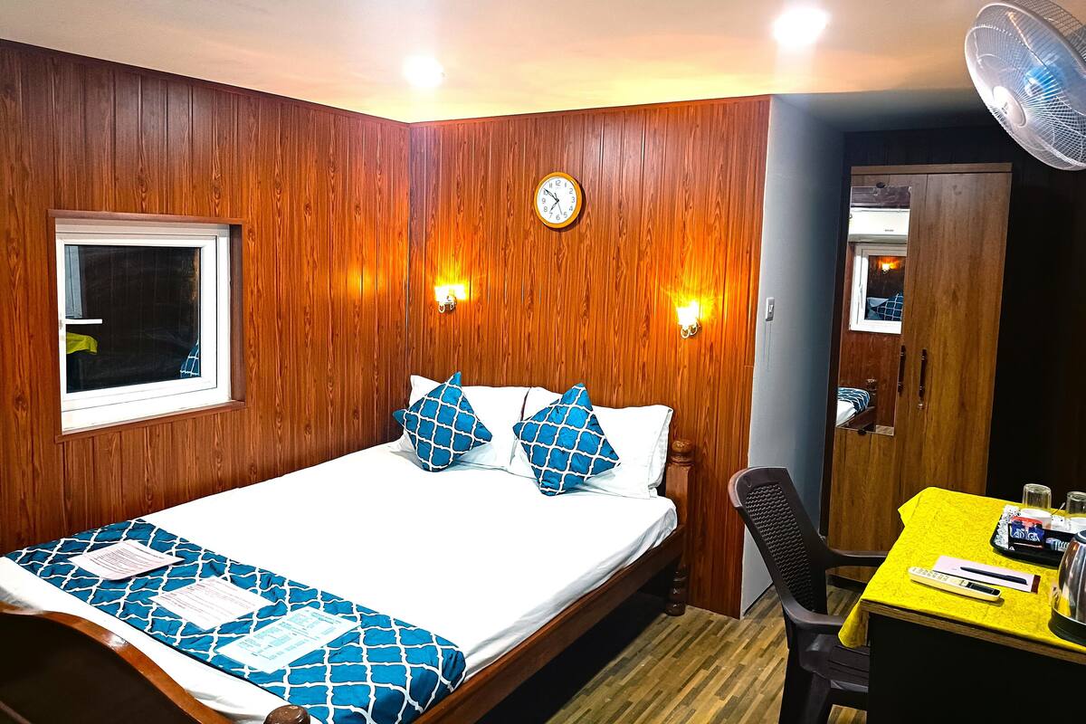 Una habitación con un panel de madera, una cama con ropa de cama de diseño azulado, un reloj y una mesa con un ventilador.
