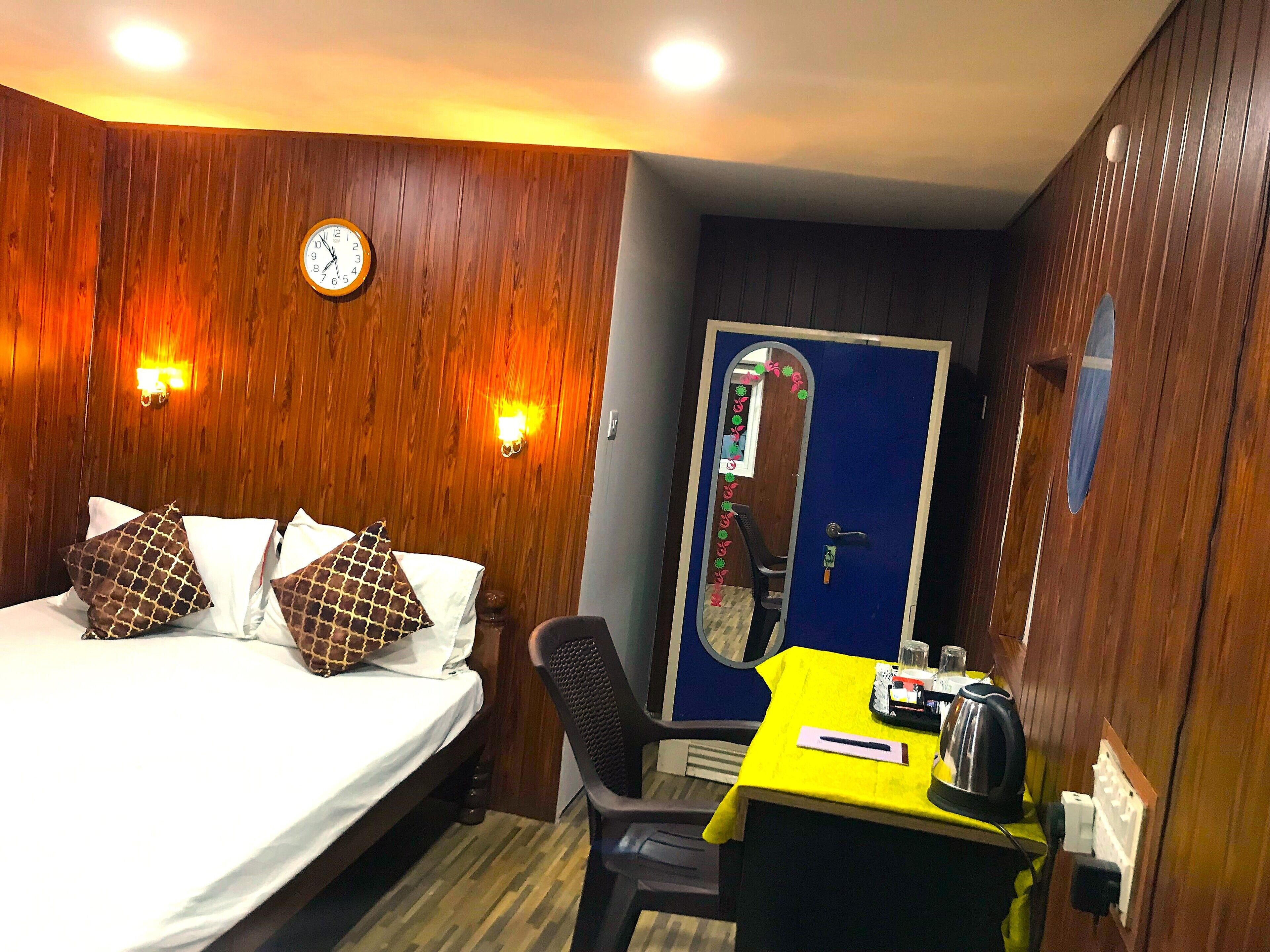 A hotel room with a bed, a desk, a chair, a clock, and a door.