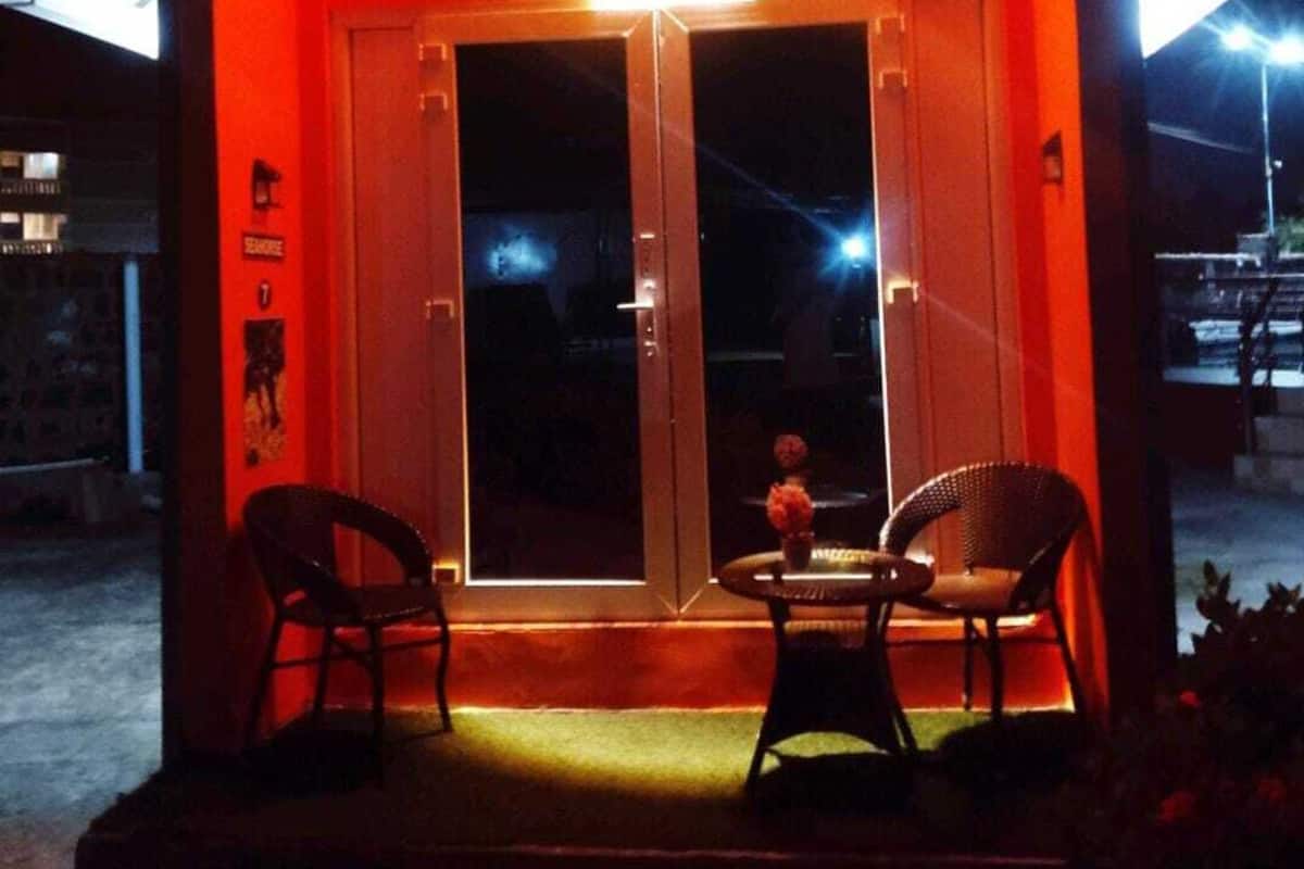 Un pequeño edificio iluminado, con techo inclinado, dos puertas de vidrio y dos sillas en el porche.