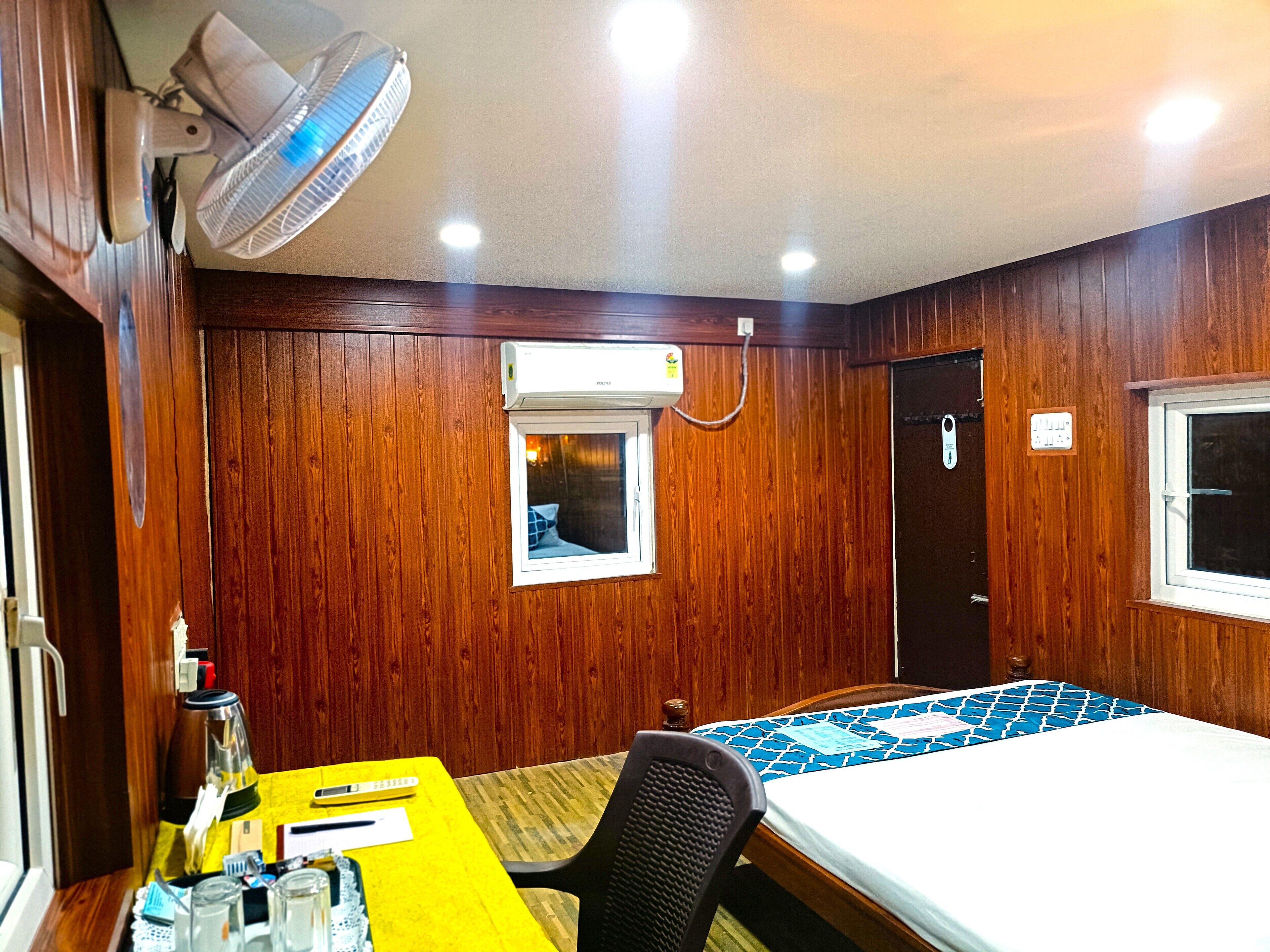 A room with wooden paneling, a bed with a blue and white patterned bedspread, a desk with a yellow surface, a black office chair, a standing fan, and an air conditioning unit.