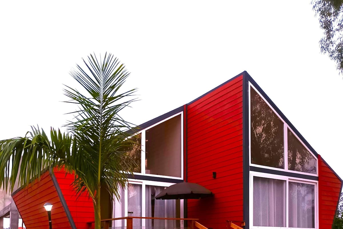 Una casa roja de dos pisos con un balcón, una escalera de madera y una palmera en primer plano.