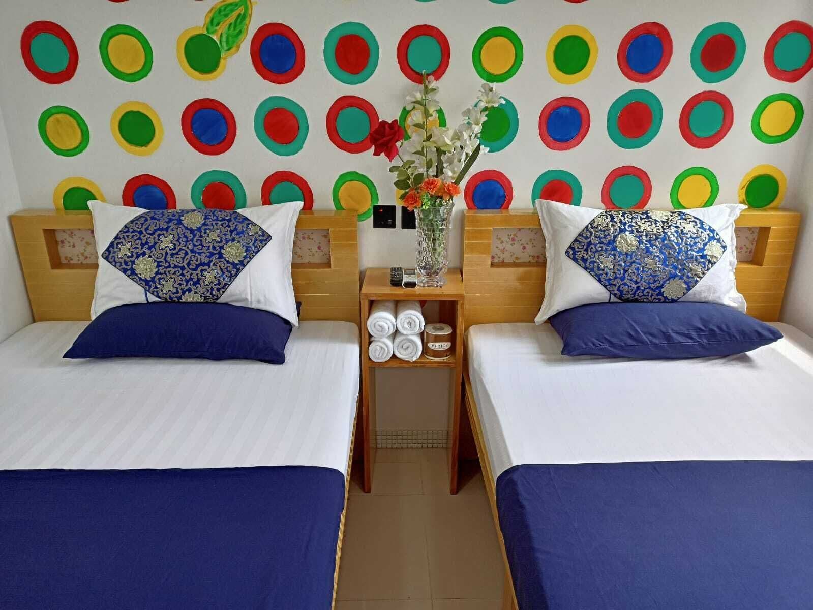 Two beds with blue bedding, a wall with colorful circular patterns, and a small table with a vase of flowers between the beds.