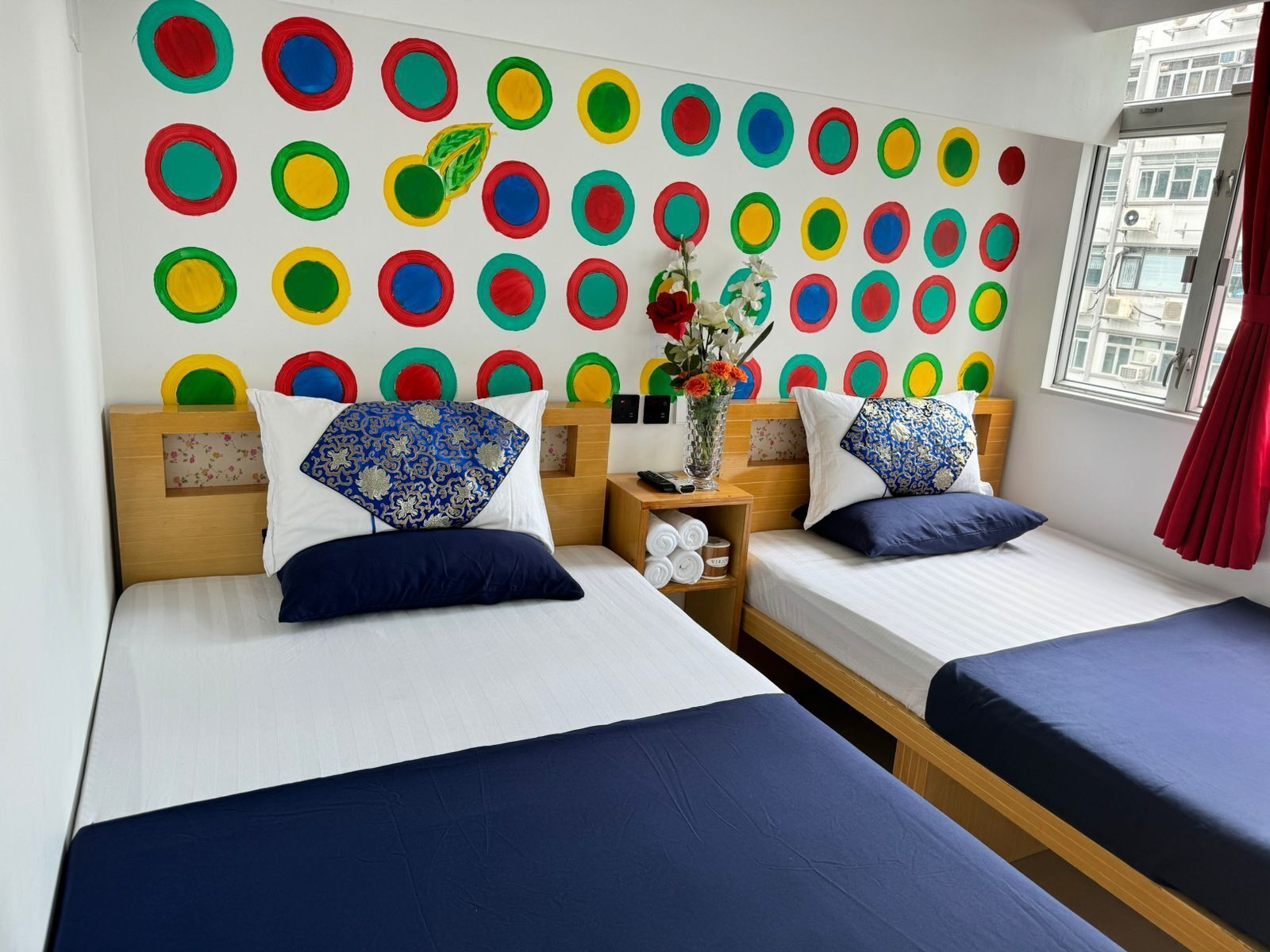 Ein Hotelzimmer mit zwei Betten, einer Wand mit bunten, kreisförmigen Mustern, einer Vase mit Blumen und einem Fenster mit Blick auf Gebäude.