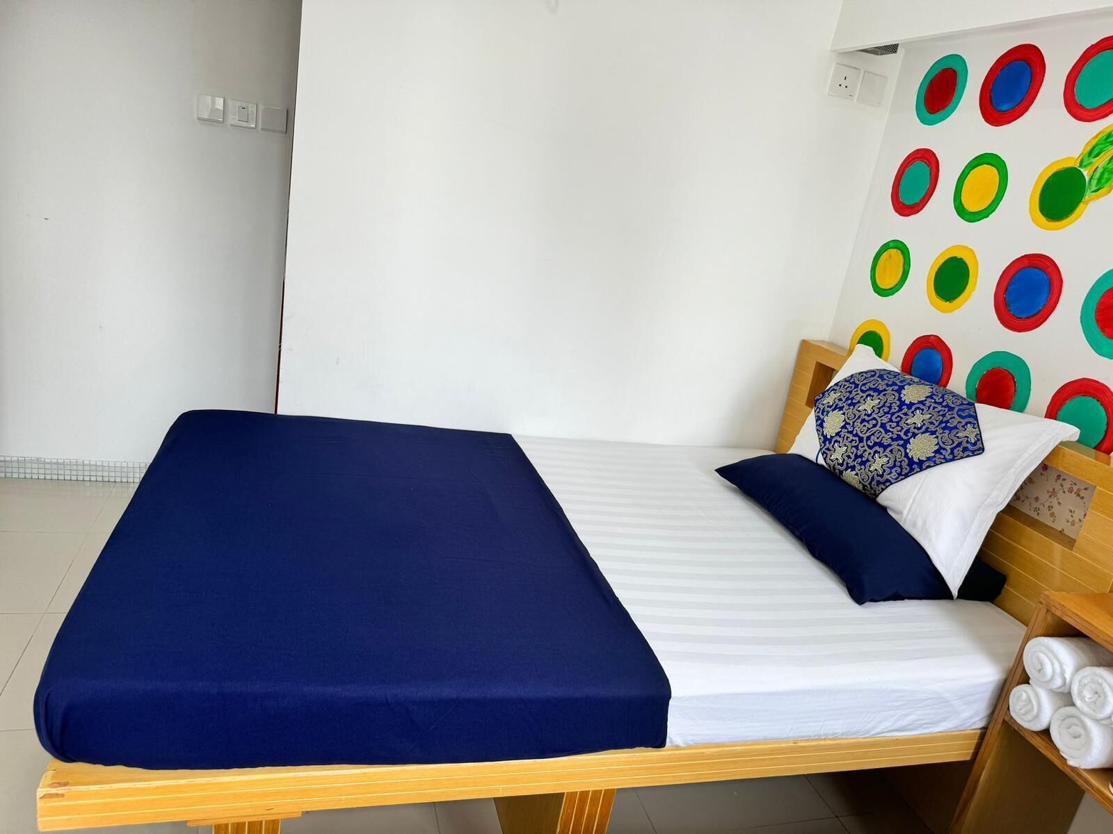 A single bed with a blue bedspread and white linens, a wooden headboard, and a wall with colorful circular patterns.
