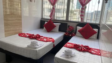 A hotel room with two beds, each with a white grid-patterned bedspread, red pillows, and a red decorative item on the bed. There is a window with pink and white striped curtains, and a wall-mounted lamp.